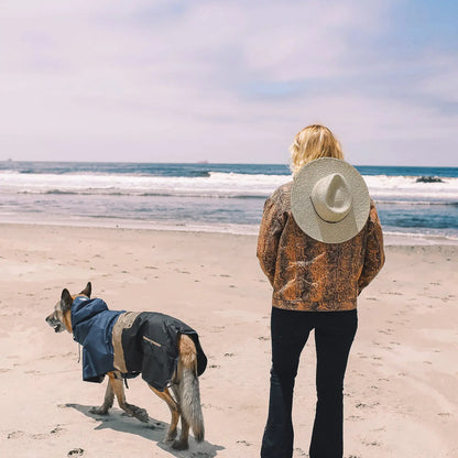 Hund im wasserdichten Baja Coastline Poncho am Strand neben seiner Bezugsperson, den Blick aufs Meer gerichtet und bereit für jedes Küstenabenteuer.