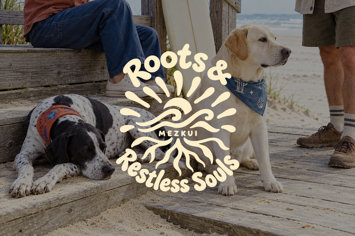 Zwei Hunde mit Halstüchern auf einer Strandpromenade mit Menschen und Surfbrett, unter einem cremefarbenen Roots & Restless Souls MEZKUI-Emblem.